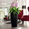 A tall black planter with a flowering plant is placed in a modern living room with red chairs and a lamp. Produktbild-Vorschau