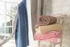 Several folded blankets are stacked on a chair next to a window with a tree outside. Produktbild-Vorschau