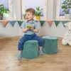 A young child is sitting on a green ottoman, holding a cup of green liquid. Another green ottoman is next to him, and a stuffed giraffe is in the background. The scene is set indoors with windows and a decorative banner. Produktbild-Vorschau