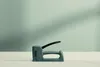A gray stapler is placed on a white surface against a light gray background with shadows. Podgląd zdjęcia produktu