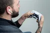 A man is holding a white and black game controller while wearing a white wireless earbud. Podgląd zdjęcia produktu