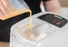 Eine Hand gießt eine gelbe Flüssigkeit aus einem Messbecher in die Unold-Nudelmaschine Nora, die ein Produktbild-Vorschau