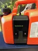 Close-up of an orange Husqvarna combi-canister with a black fuel cap and a black compartment for tools or accessories. The background shows a blurred image of trees and foliage. Voir les images du produit