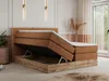 A bedroom scene featuring a large boxspring bed with its storage compartment open, showcasing the mattress and wooden frame. Produktbild-Vorschau
