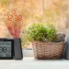 A projection alarm clock with weather forecast is placed on a windowsill next to a basket of green flowers, with raindrops visible on the window. Produktbild-Vorschau