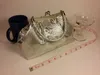 A silver clutch purse is displayed next to a blue mug, a champagne coupe, and a measuring tape. Podgląd zdjęcia produktu