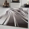 A modern abstract wave design rug in shades of gray and white is shown on a light gray floor, with a fireplace and stacked logs in the background. Náhľad obrázku produktu