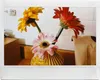 Three gerbera daisies in a yellow vase on a wooden surface with a white tiled background. Produktbild-Vorschau