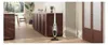 A white stick vacuum cleaner stands upright on a wooden floor in a modern kitchen setting, with dark wood cabinets and a large window in the background. Voir les images du produit
