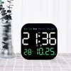 A digital clock is displayed on a table next to a vase with plants. Produktbild-Vorschau