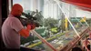 A person aims a sniper rifle from a window overlooking a race track with skyscrapers in the background. Náhľad obrázku produktu