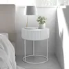 A white round bedside table with a plant and a lamp on it, next to a bed. Produktbild-Vorschau