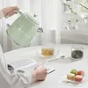 A person is pouring hot water from a green electric kettle into a clear glass cup of tea, with macarons and tea leaves on the table. Voir les images du produit