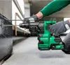 A person is using a green and black cordless hammer drill to drill into a dark gray wall. Voir les images du produit