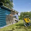 A man is painting a wooden shed with a spray gun. A yellow paint sprayer is next to him. Náhled obrázku produktu