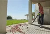A woman is using a leaf blower on a wooden deck with debris scattered on it. The background shows a green lawn and a clear sky. Produktbild-Vorschau