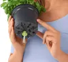 A person is holding a plant pot upside down, showing the bottom with a self-watering system. Náhled obrázku produktu