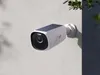 A white security camera with a solar panel on top is mounted on a textured gray wall. A shadow of the camera and a branch with green leaves are visible on the wall. Náhľad obrázku produktu