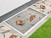 A metal grate covering a basement window well, with fallen leaves scattered around it and on top of it. The grate is set into a stone patio next to a patch of green grass. Produktbild-Vorschau