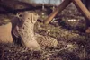 A pair of beige tactical boots resting on the ground in an outdoor setting. Produktbild-Vorschau