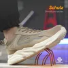 A person wearing a tan sneaker-style work shoe is stepping on a metal object with sharp points, demonstrating the shoe's protective features. The background is blurred and shows construction-like elements. Produktbild-Vorschau