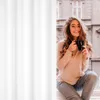 A young woman is sitting by a window, holding a mug and smiling. Produktbild-Vorschau