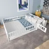 A white wooden children's bed with a blue and white patterned quilt and pillows, set up in a room with light blue walls and a grey carpet. Produktbild-Vorschau