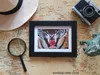 A flat lay composition featuring travel-related items on a map background, including a framed picture of a tiger, a camera, a magnifying glass, and a hat. Produktbild-Vorschau