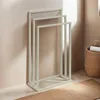 A beige metal towel rack with three bars stands against a light-colored wall, with a woven basket and a rug visible. Produktbild-Vorschau