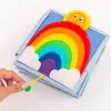 A close-up shot of a baby's quiet book with a rainbow and sun design, held by a person's hand. Produktbild-Vorschau