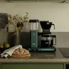 A Moccacoffee maker sits on a kitchen counter next to a loaf of bread and some fruit. Náhled obrázku produktu