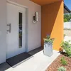 A modern house entrance with a white door, a doormat, and a planter with flowers. Produktbild-Vorschau