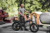 A man stands next to a black electric bicycle in an outdoor setting with industrial equipment and a large rock in the background. Produktbild-Vorschau