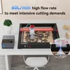 A laser engraving machine with an air assist system is shown on a desk, actively engraving a wooden board with a dragon design. There are also small wooden models of a car and a house in the background. Produktbild-Vorschau