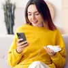 A woman is sitting on a couch, holding a smartphone and a white power bank, with a charging cable connecting them. Náhled obrázku produktu