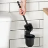 A hand holding a black toilet brush over its black holder, mounted on a white tiled wall next to a toilet. Náhled obrázku produktu