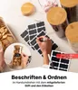 Glasvorratsgläser mit Bambusdeckeln, Tafeletiketten und einem weißen Kreidestift auf einer weißen Marmoroberfläche. Eine Hand schreibt 'Cookies' auf ein Etikett Voir les images du produit