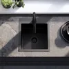 A black kitchen sink with a black faucet is shown from a top-down view, placed on a grey stone countertop. There are two small potted plants on a white shelf in the background. Náhled obrázku produktu
