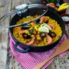 A paella dish in a pan on a wooden table, with a checkered cloth and a wooden spoon. Podgląd zdjęcia produktu