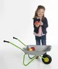 A young girl holding apples stands next to a toy wheelbarrow. Produktbild-Vorschau