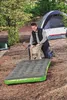A man is kneeling on the ground in a wooded area, using a hand pump to inflate an air mattress. A tent is visible in the background. Podgląd zdjęcia produktu
