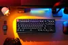 A desk setup with a keyboard, mouse, headphones, watch, and game controller, illuminated by colorful LED lights. Produktbild-Vorschau