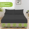 A bed with dark gray fitted sheets and two pillows, set in a bedroom with a window and a small side table with a plant and a cup. Produktbild-Vorschau