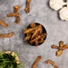 A flat lay of dried chicken necks on a textured gray surface, with a small potted plant and two rope toys in the background. Produktbild-Vorschau