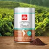 A can of Illy coffee is placed on a wooden surface in front of a blurred background of coffee plants. Produktbild-Vorschau