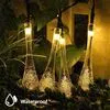 A string of solar-powered fairy lights with teardrop-shaped crystal bulbs hanging among greenery. Produktbild-Vorschau