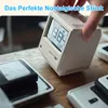 A hand holding a retro-style electronic alarm clock that resembles an old computer monitor, placed on a wooden desk with other electronic devices. Produktbild-Vorschau