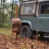 A rusty outdoor fireplace is placed on the ground in a forest setting next to a green off-road vehicle. Anteprima dell’immagine prodotto