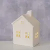 A white ceramic house-shaped decoration with illuminated windows sits on a white surface against a mottled gray background. Produktbild-Vorschau