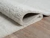 A close-up view of a cream-colored shaggy rug, partially rolled, revealing its backing and the wooden floor beneath. Produktbild-Vorschau